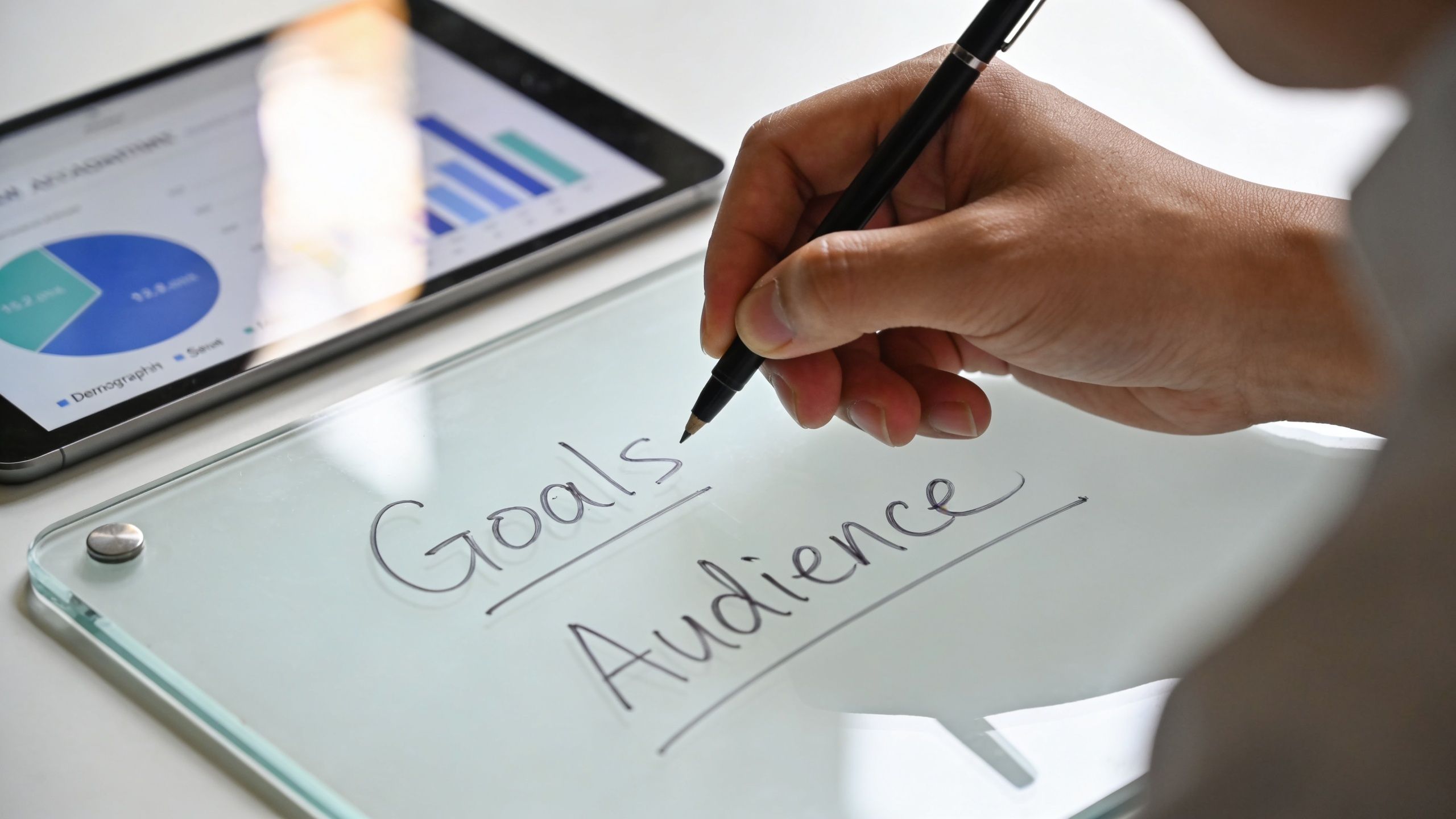 A professional writing the words Goals and Audience on a glass desktop board during a business meeting.
